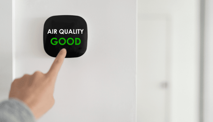 Finger pointing to an indoor air quality monitor displaying "Air Quality Good" mounted on a white wall
